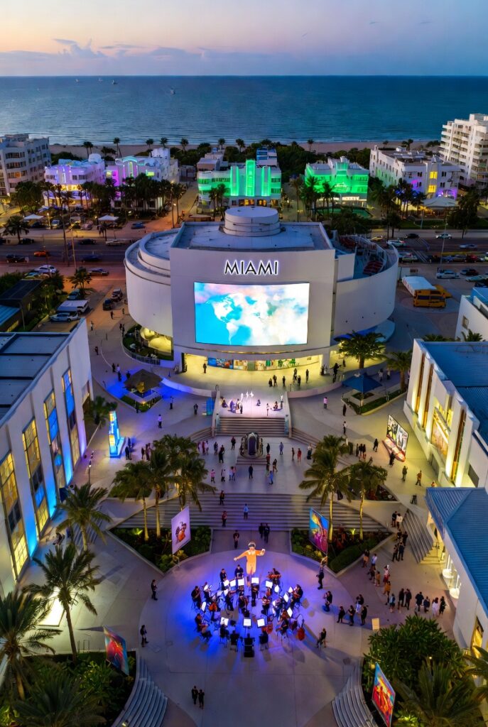 Aerial view of Miami’s cultural district at sunset, featuring an illuminated building that reads “Miami,” an outdoor musical event, palm trees, and the Atlantic Ocean in the background